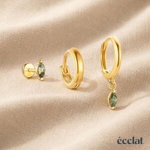 May include: Three gold-colored earrings on a beige surface. One is a stud with a green gemstone, another is a small hoop, and the third is a hoop with a dangling green gemstone. The brand name "&eacute;clat" is at the bottom.