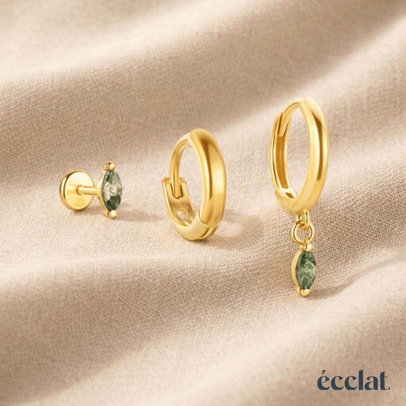 May include: Three gold-colored earrings on a beige surface. One is a stud with a green gemstone, another is a small hoop, and the third is a hoop with a dangling green gemstone. The brand name "&eacute;clat" is at the bottom.