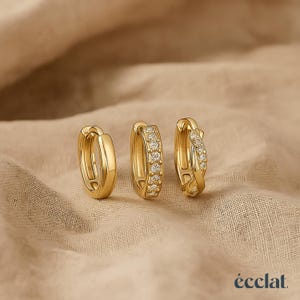May include: Three gold-colored hoop earrings. One is a plain, polished gold. The second has a row of small, clear gemstones. The third has a twisted design with clear gemstones. The earrings are on a neutral-colored fabric.