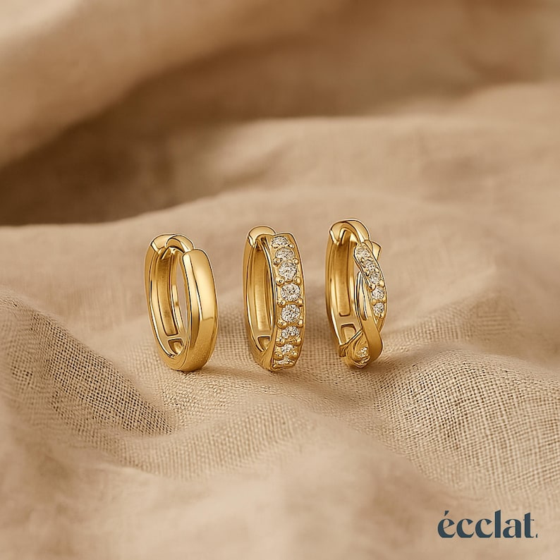 May include: Three gold-colored hoop earrings. One is a plain, polished gold. The second has a row of small, clear gemstones. The third has a twisted design with clear gemstones. The earrings are on a neutral-colored fabric.