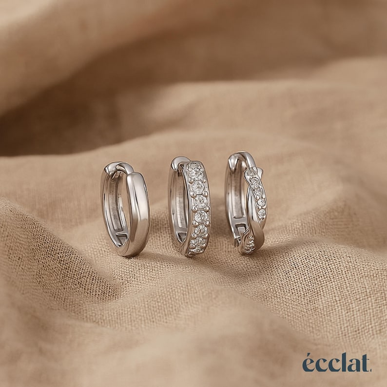 May include: Three silver hoop earrings. One is a simple, polished design. The second features a row of small, clear stones. The third has a twisted design with clear stones. The earrings are displayed on a neutral, textured surface.