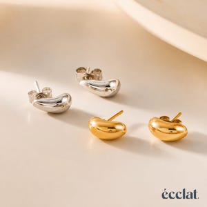 May include: Two pairs of stud earrings in a studio setting. One pair is silver, the other is gold. The earrings have a smooth, bean-like shape. The brand name "&eacute;clat" is in the bottom right corner.