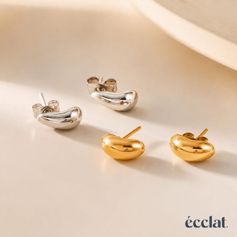 May include: Two pairs of stud earrings in a studio setting. One pair is silver, the other is gold. The earrings have a smooth, bean-like shape. The brand name "&eacute;clat" is in the bottom right corner.