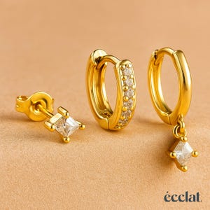 May include: A set of three gold-colored earrings. One is a stud with a square, clear stone. The second is a small hoop with clear stones. The third is a hoop with a square, clear stone pendant. The brand name "éclat" is at the bottom.