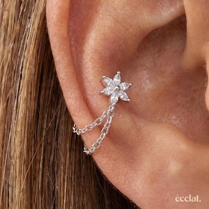 May include: A silver-toned ear cuff featuring a flower-shaped charm with six clear, marquise-cut stones arranged in a petal pattern. Two delicate silver chains extend from the base of the flower, connecting to a smooth, curved metal band.