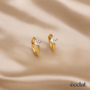 Könnte beinhalten: Ein Paar goldfarbene Creolen, jeweils mit einem klaren, quadratisch geschliffenen Edelstein. Die Ohrringe werden auf einer beigen, satinartigen Oberfläche präsentiert. Der Markenname "éclat" ist unten rechts aufgedruckt.