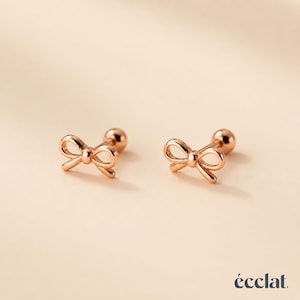 Può includere: Un paio di orecchini a lobo in oro rosa a forma di delicati fiocchi con chiusura a sfera. Gli orecchini presentano una finitura metallica lucida e un design semplice con anelli aperti. La parola "écclat." è stampata nell'angolo in basso a destra.