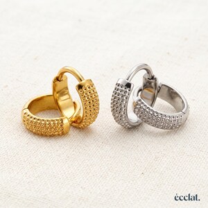May include: A pair of gold-toned hoop earrings and a pair of silver-toned hoop earrings. Each hoop features a textured surface with a grid of small, raised circular bumps. The word "écclat." is printed in the bottom right corner of the image.