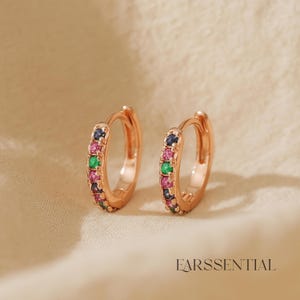Pode incluir: Um par de brincos de argola em ouro rosa. Cada brinco é adornado com uma fileira de pedras preciosas coloridas em tons de rosa, verde e azul. Os brincos são exibidos em um fundo neutro, com a palavra "EARSSENTIAL" visível.