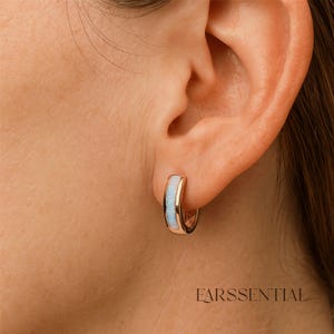 Peut inclure: Boucle d'oreille cr&eacute;ole en or rose avec incrustation bleu clair. La boucle d'oreille est de petite taille, de forme incurv&eacute;e, avec une finition m&eacute;tallique polie et une pierre bleu clair contrastante. Le mot "EARSSENTIAL" est imprim&eacute; en bas de l'image.
