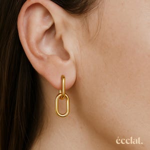 May include: Gold-coloured chain link earrings. The earrings are a bright, shiny gold and are attached to the ear. The earrings are a simple, yet elegant design. The word "éclat" is in the bottom right corner.