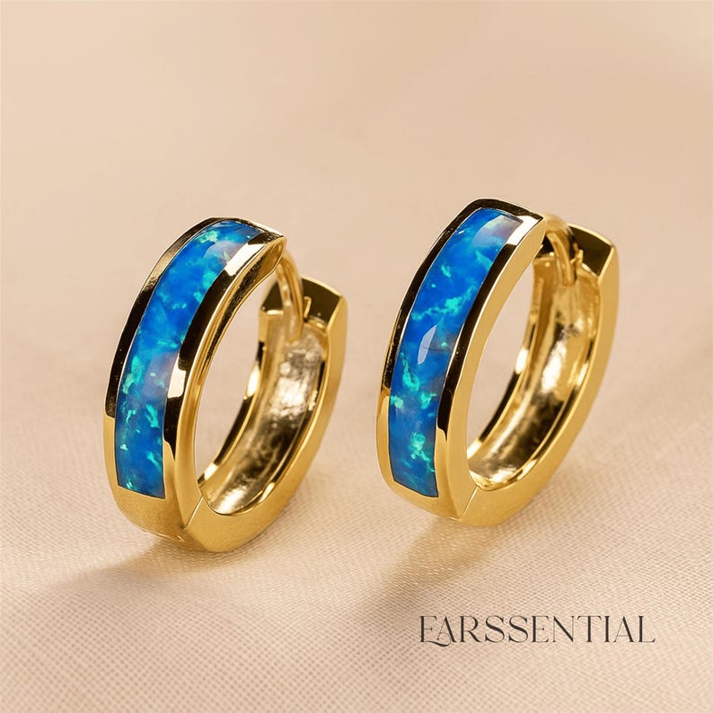 Peut inclure: Deux boucles d'oreilles cr&eacute;oles dor&eacute;es avec une incrustation d'opale bleue. Les boucles d'oreilles ont une finition polie et un design classique. Le texte "EARSSENTIAL" est visible en bas.