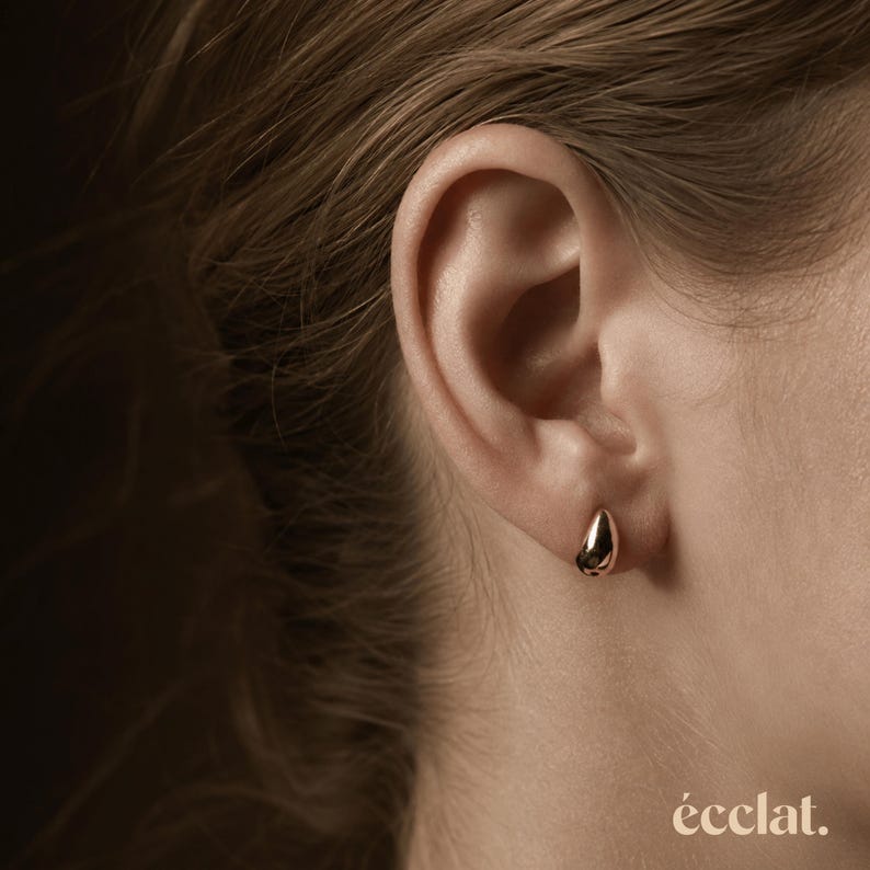 May include: Close-up of a gold teardrop earring on an ear. The earring is smooth and reflective, catching the light. The word "&eacute;clat" is printed in a small font at the bottom right corner of the image.