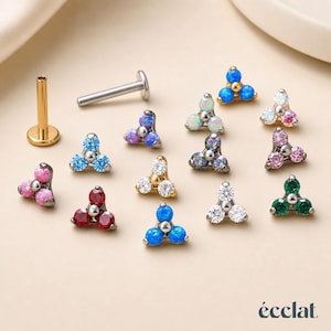 May include: A selection of ear studs with a three-stone design in various colours, including blue, pink, green, and white. The image also shows a gold and silver post. The word "éclat" is at the bottom.