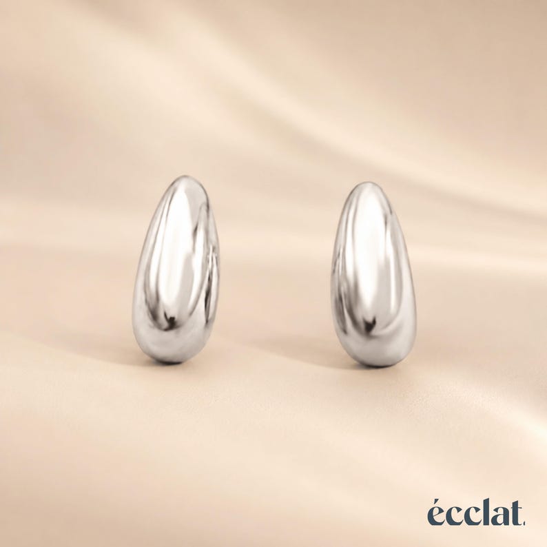 May include: A pair of polished silver teardrop-shaped earrings. The earrings have a smooth, reflective surface and are displayed against a neutral, cream-colored background. The word "&eacute;clat" is printed in the bottom right corner.