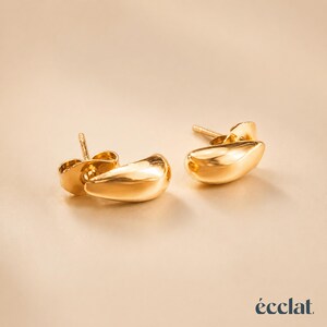 May include: A pair of gold-colored stud earrings with a teardrop design. The earrings have a smooth, polished surface and a post backing. The earrings are displayed on a neutral background. The word "&eacute;clat" is in the bottom right corner.