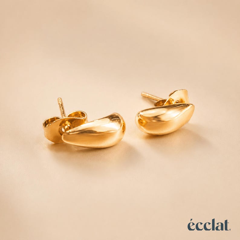 May include: A pair of gold-colored stud earrings with a teardrop design. The earrings have a smooth, polished surface and a post backing. The earrings are displayed on a neutral background. The word "&eacute;clat" is in the bottom right corner.