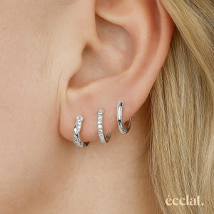 May include: Three silver hoop earrings of varying designs are displayed on an ear. The first earring has a twisted design, the second is encrusted with small stones, and the third is a plain, polished hoop. The word "eclat" is in the bottom right corner.