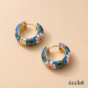 Può includere: Un paio di orecchini a cerchio color oro con una base in smalto blu brillante e un motivo floreale di petali bianchi e rosa con foglie verdi. Gli orecchini presentano una chiusura a cerniera e la parola éclat è stampata in basso a destra.