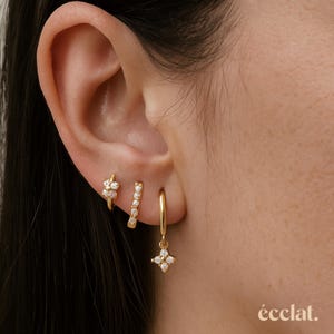 May include: A close-up of an ear adorned with three gold earrings. The earrings include a small flower-shaped hoop, a vertical bar with small stones, and a hoop with a dangling four-petal charm. The brand name "éclat" is visible.