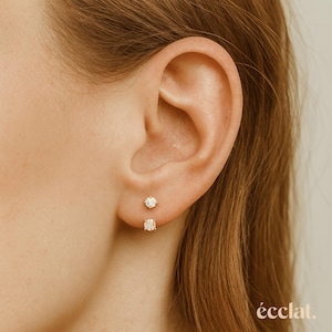 May include: Close-up of an ear with two small diamond stud earrings. The top earring is round, and the bottom earring is square. The earrings are set in gold. The ear is framed by light brown hair.