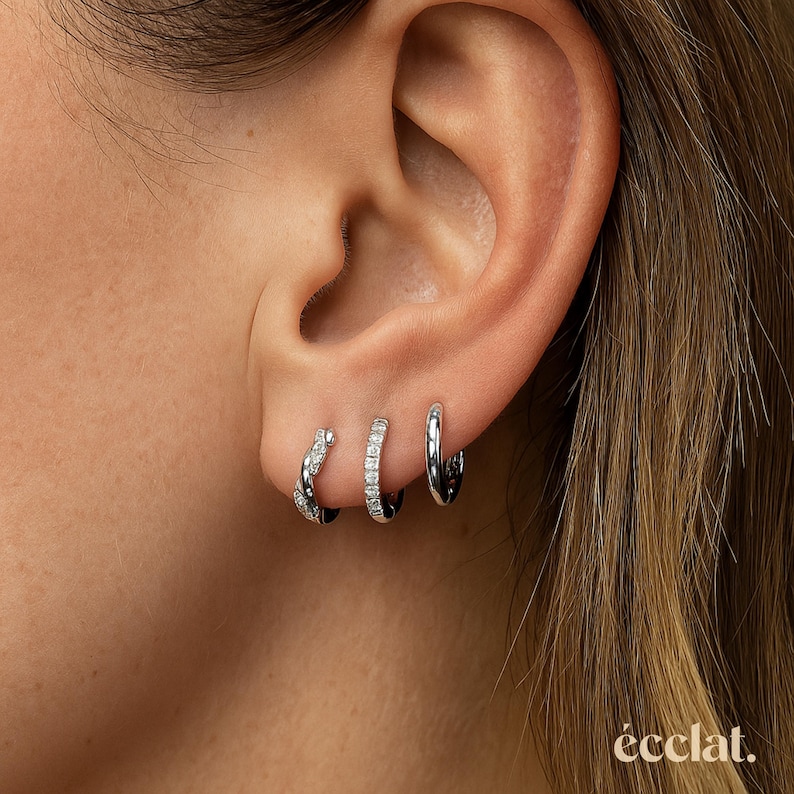 May include: Three silver hoop earrings of varying sizes are displayed on an ear. The smallest hoop is encrusted with small, clear stones. The middle hoop is also encrusted with clear stones. The largest hoop is a plain, polished silver.