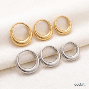 Peut inclure: Une collection de six boucles d'oreilles créoles disposées sur deux rangées. La rangée supérieure comprend trois créoles dorées, et la rangée inférieure trois créoles argentées. Chaque boucle présente une surface lisse, arrondie et polie.