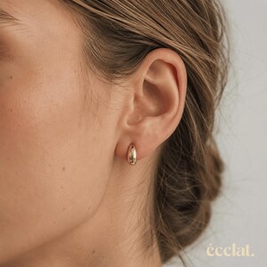 May include: A close-up of a gold-colored hoop earring worn on an ear. The earring is a small, rounded shape. The background is a neutral color.