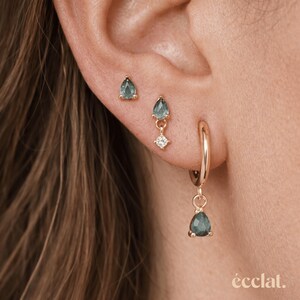 May include: Close-up of three gold earrings with teal teardrop-shaped gemstones. One is a stud, another dangles with a small diamond, and the third hangs from a hoop earring. The word "&eacute;clat" is at the bottom.