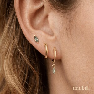 May include: Close-up of an ear with three gold earrings. One is a small stud with a green gemstone, and two are gold hoops. One hoop has a dangling green gemstone. The word "eclat" is in the bottom right corner.