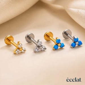 Op de afbeelding: Vier sieraden, elk met een platte schijfbasis en een staaf. De sieraden hebben een driehoekig ontwerp met drie stenen. Twee zijn goudkleurig met heldere stenen, één is zilverkleurig met heldere stenen en twee zijn goudkleurig met blauwe stenen.