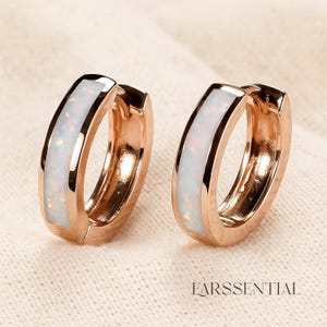 Peut inclure: Deux boucles d'oreilles cr&eacute;oles en or rose avec un insert en opale blanche. Les boucles d'oreilles ont une finition polie et une fermeture &agrave; charni&egrave;re. Le nom de la marque "EARSSENTIAL" est imprim&eacute; en bas de l'image.