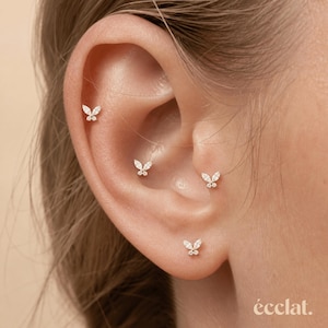 May include: Close-up of an ear adorned with four small, gold butterfly earrings. Each earring features a delicate butterfly design with sparkling accents. The earrings are positioned at different points on the ear, showcasing their placement and design. The word "éclat" is at the bottom right.