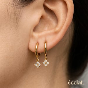 May include: Gold hoop earrings with a four-petal flower charm, each petal set with small, clear gemstones. The earrings are shown on an ear, with the brand name "éclat." in the lower right corner.