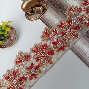 Pode incluir: Acabamento floral bordado com flores vermelhas e douradas e detalhes de contas. O acabamento tem uma base transparente esbranquiçada e é adornado com lantejoulas. Uma estatueta de elefante dourado também é visível.