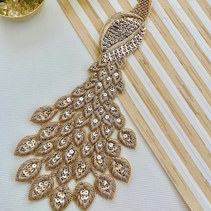 May include: A gold sequined peacock applique with a detailed design. The peacock has a long, flowing tail and is embellished with small, shiny sequins. The bird's body is made of woven gold threads. The applique is suitable for embellishing clothing or home decor.