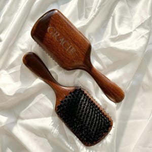 以下が含まれることがあります： 濃い色の毛とカーブした柄の木製ヘアブラシ2本。上のブラシには「GRACIE」という名前が刻まれています。ブラシは濃い茶色で、白い背景に置かれています。