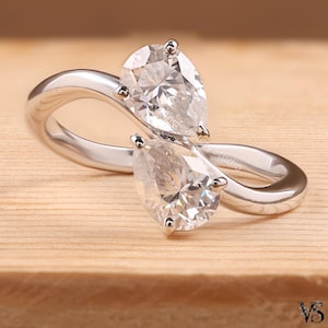 May include: A bypass ring crafted from polished silver-toned metal, featuring two pear-cut clear stones set in a continuous, flowing band design. The stones are positioned at opposite ends of the open, overlapping structure of the ring.
