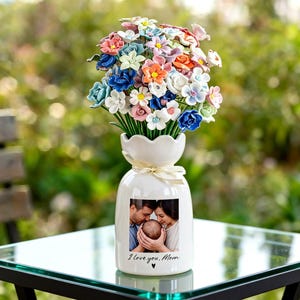 May include: A white ceramic vase with a photo and the text "I love you, Mom" holds a bouquet of colorful artificial flowers. The flowers include blue, pink, orange, and white blossoms. A ribbon is tied around the vase's neck.