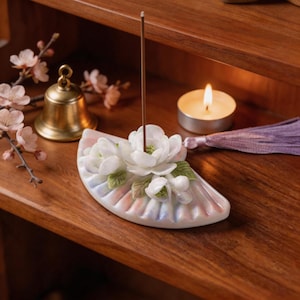 May include: A ceramic incense holder with white floral accents and a brown incense stick. The holder is fan-shaped with pastel pink and blue hues. A small gold bell, a lit candle, and a purple tassel are also visible.