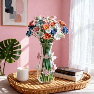 Puede incluir: Un jarrón de cristal lleno de coloridas flores de cerámica, incluyendo flores azules, naranjas y blancas. El jarrón está adornado con mariposas y se encuentra sobre una bandeja tejida con una vela encendida y libros. El fondo es una pared rosa.