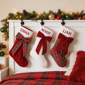 Pode incluir: Três meias de Natal penduradas em ganchos pretos. A primeira é vermelha com um laço xadrez e o nome EMMA. A segunda é branca com um laço de veludo vermelho e o nome NOAH. A terceira é xadrez com um laço de veludo vermelho e o nome LIAM.