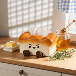 May include: A rectangular tissue box cover shaped like a loaf of bread with a golden-brown crust and a white center. The front features a cartoon face with two black eyes, a small heart-shaped mouth, and two small brown feet at the base.