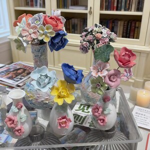 May include: A collection of ceramic vases and floral arrangements. The vases are white, with pastel-colored flowers in shades of pink, blue, yellow, and orange. Some vases are tall and cylindrical, while others are small and egg-shaped.
