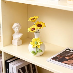 Puede incluir: Un jarrón de cerámica blanca con detalles de girasoles amarillos contiene tres girasoles con centros marrones. Un girasol decorativo está adherido al jarrón. Un busto de una persona está en el estante.