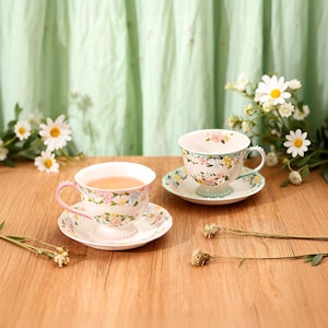 May include: Two floral teacups with matching saucers are displayed on a wooden surface. One cup is filled with tea. The cups and saucers are white with pink, yellow, and blue floral patterns. The teacups have pink handles and are set against a green backdrop.