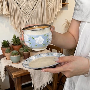 May include: A ceramic tea cup and matching saucer set featuring a light blue glaze decorated with white flowers and green leaves. The cup has a cream-colored rim and a scalloped edge, while the saucer displays a matching floral pattern.