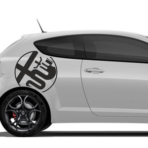 Pode incluir: Um carro hatchback branco com um decalque de logotipo preto na lateral. O decalque é um design circular com um emblema estilizado. O carro tem rodas pretas e vidros fumados. O carro está estacionado em uma superfície.