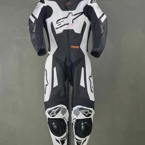 May include: A black and white motorcycle racing suit with the word "MATRIX" in orange. The suit has a full-body design with protective padding on the knees and elbows. The suit is hanging on a hanger against a gray background.