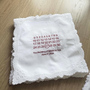 May include: White handkerchief with a delicate lace trim. Embroidered in burgundy are numbers forming a calendar, with the 27 circled. Text reads "The Wedding of KENNY&TINA June 27, 2026."